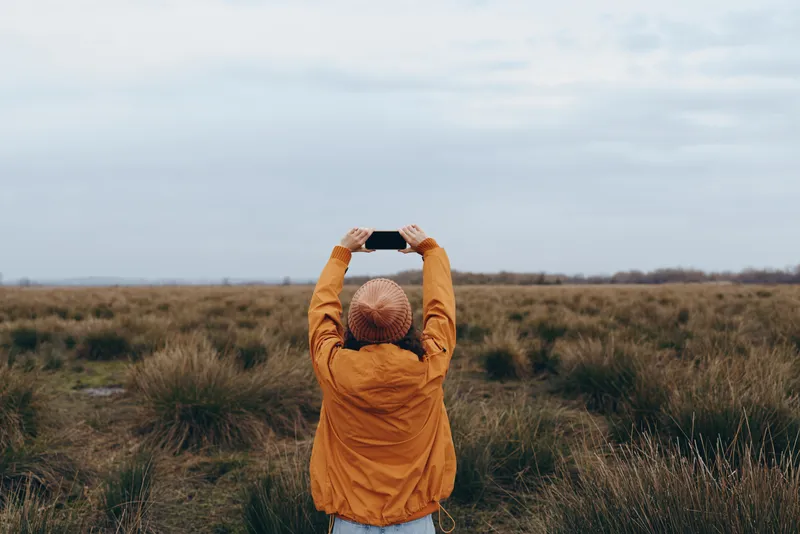 Lifestyle woman outdoors in orange coat stands in field of grass, holding a hat above head, back view, cloudy sky, wide landscape, calm windy mood