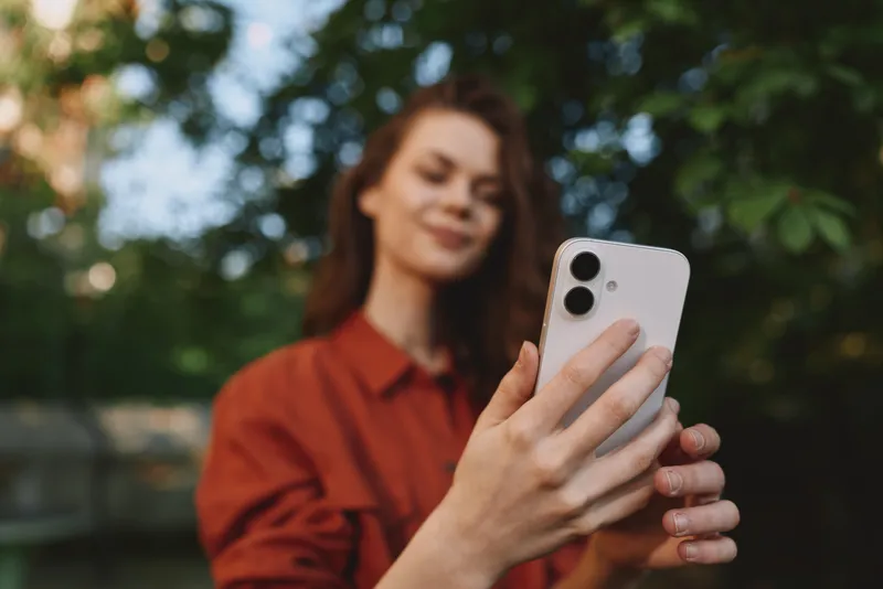 Young woman lifestyle portrait taking a selfie outdoors in a red shirt, smiling softly while holding a smartphone, surrounded by lush green trees and warm daylight