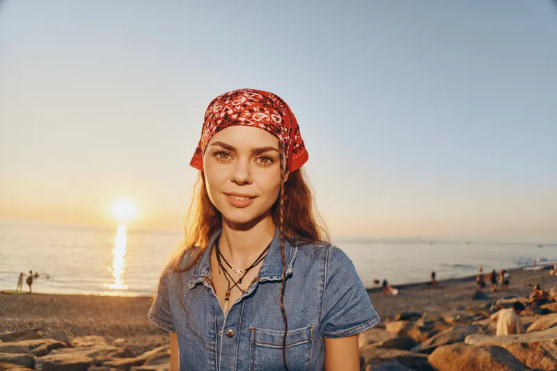 lifestyle woman in modern boho-western denim street style with a red bandana and layered necklaces at sunset beach, film color warm tones