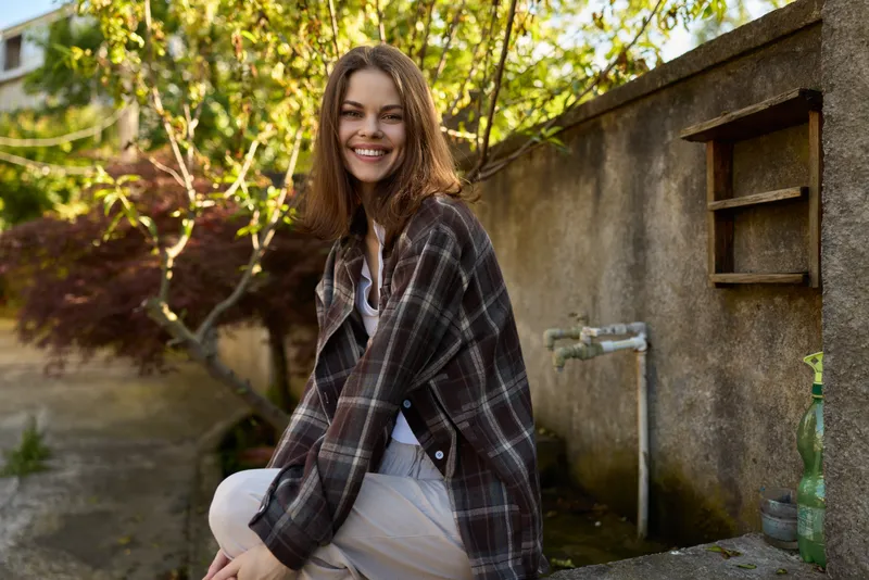 A cheerful woman sits outdoors in daytime, wearing a plaid jacket and casual outfit. A relaxed, candid portrait capturing lifestyle, warmth, and approachable fashion