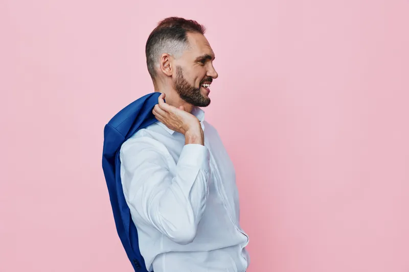 A professional man stands in profile against a pink backdrop, in a light shirt with a jacket over his shoulder, exuding confidence, optimism, and a friendly business vibe.