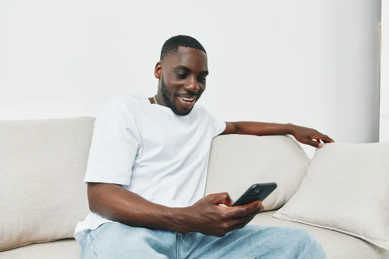 Young Black man in casual attire smiling while using smartphone, sitting on a light colored sofa, expressing joy and connection in a modern interior