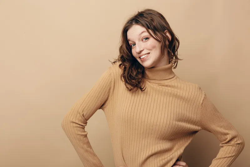 Smiling young woman with curly brown hair wearing beige knitted sweater poses confidently against beige background. Concept of casual style, happiness, and natural beauty in studio portrait.