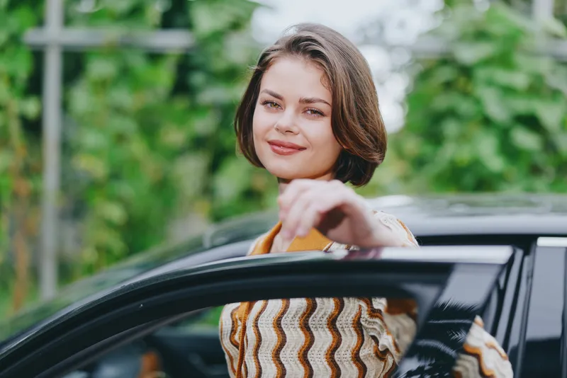 Portrait of a woman leaning from a car window, smiling at the camera, in a striped sweater and natural daylight.