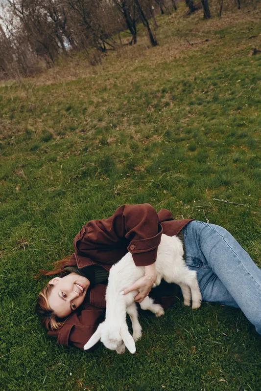 Playful moment of a woman outdoors, lying on grass, cuddling a white goat kid, warm clothing, sunlit scene, relaxed mood, gentle breeze, natural colors, simple countryside setting