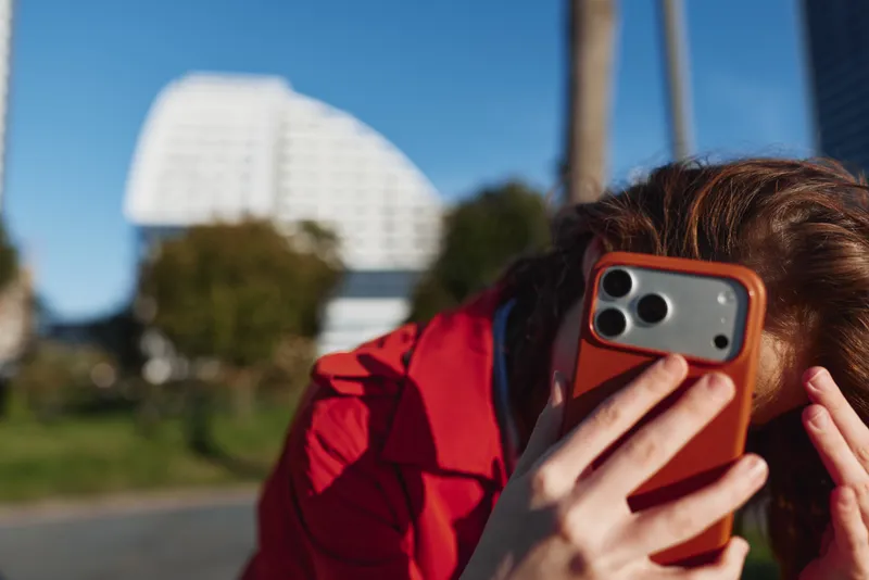 Young woman with brunette hair speaks on an orange phone case outdoors, wearing a red jacket, in a sunny park with a tall building in the background.