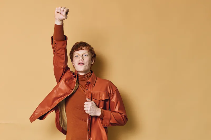 Young man standing with raised fist in burnt orange jacket and turtleneck against plain background, symbolizing confidence, strength, and empowerment gesture.