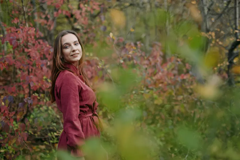 Portrait of a young woman in a red coat among autumn foliage, smiling with relaxed posture and soft gaze. The frame emphasizes a genuine, truthful outdoor moment for natural portrait storytelling.