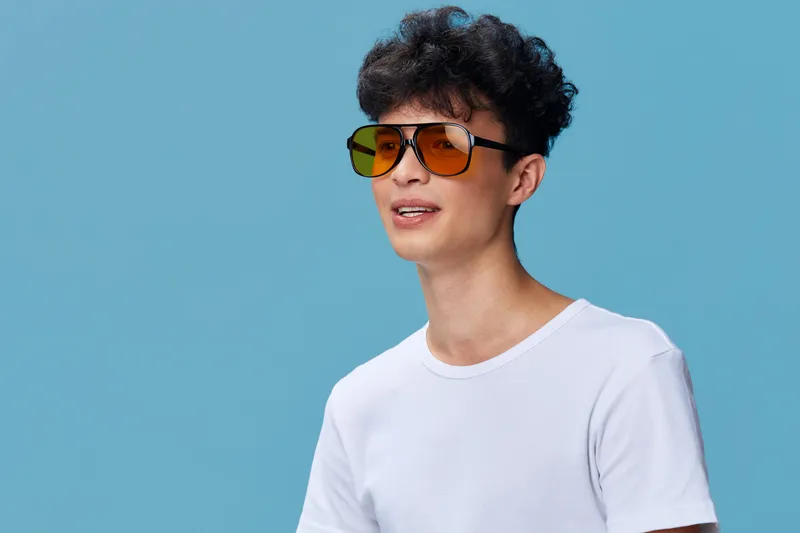 Young man with curly hair wearing yellow sunglasses and a white t-shirt smiles against a blue background, showcasing casual style and modern fashion accessories.