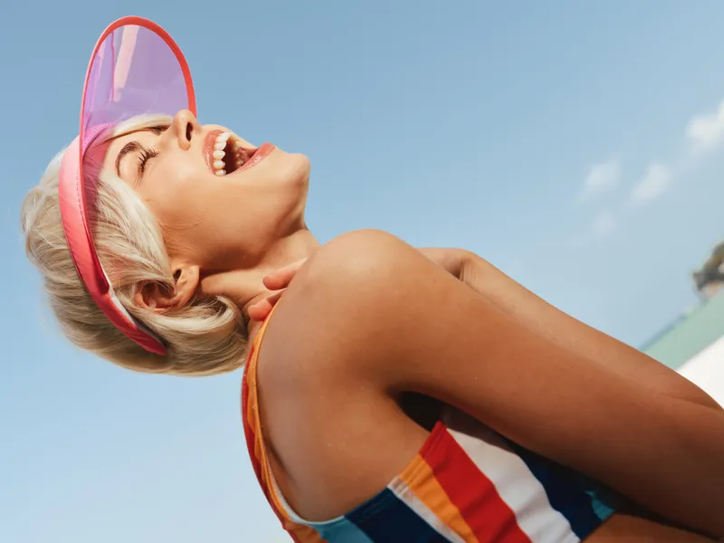 Bright portrait of a joyful woman wearing a pink visor and colorful striped swimsuit, laughing softly while looking up, against a clear blue sky.