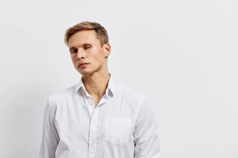 Thoughtful young man with short brown hair in white shirt looking away, standing against plain white background. Studio portrait. People, lifestyle, contemplation, male, casual, neutral tone, fashion 