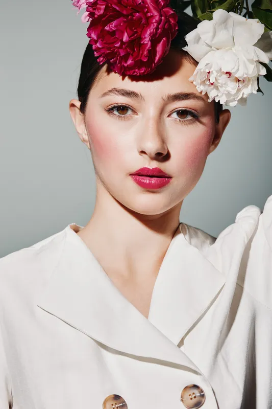 Creative beauty portrait of a woman with bold makeup wearing large red and white flowers in her hair. She wears a white blazer, posing on a light gray background.