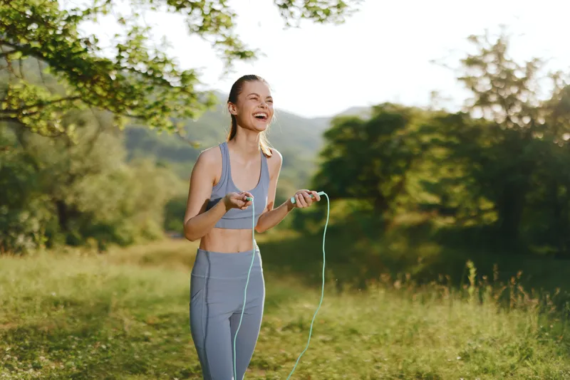 Young woman laughing while skipping rope in nature, showcasing fitness and joy in a sunny outdoor setting, perfect for healthy lifestyle themes