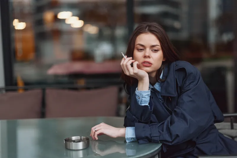 Young woman in dark coat sitting at outdoor cafe table smoking cigarette. Urban setting with glass table and blurred background. Concept of contemplation and relaxation.