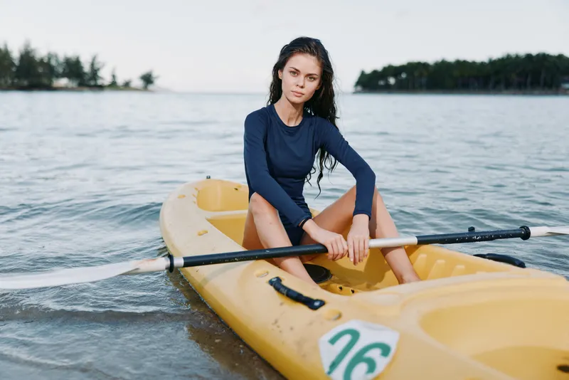 A young woman sits in a yellow kayak on calm water, wearing a dark blue long sleeve top, ready to paddle, with gentle waves and a distant tree line.