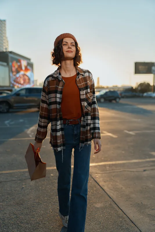 woman walking golden hour candid urban paper beanie plaid smile captures candid lifestyle and authenticity in a city parking lot; emotional storytelling, mindful living and golden hour glow with
