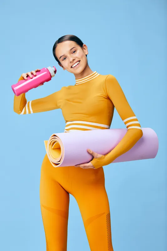 Fitness woman in bright orange athletic wear holds a pink water bottle and a rolled yoga mat against a blue backdrop, expressing energy, readiness, and active lifestyle.