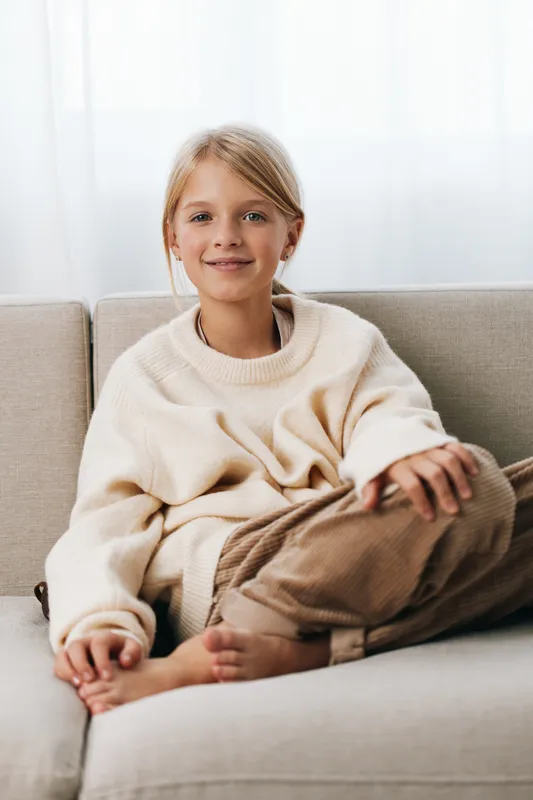 A smiling young girl sits relaxed on a soft sofa, wearing a cream cozy sweater and brown pants, creating a warm, inviting domestic portrait.