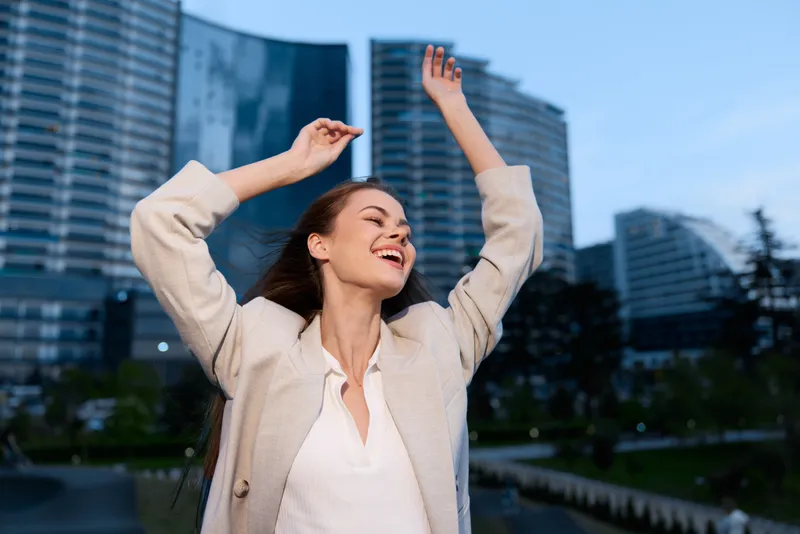 Confident woman in business attire joyfully celebrating success against a modern cityscape backdrop, showcasing empowerment and ambition.