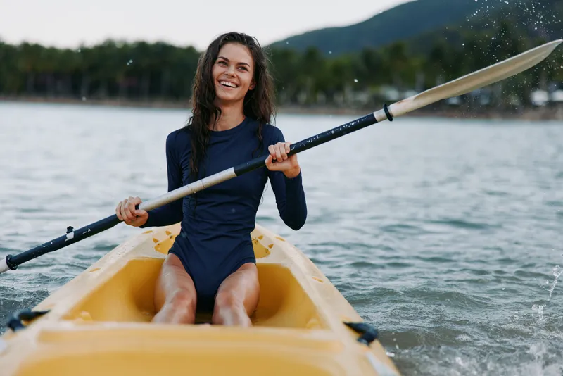 Smiling woman paddles a yellow kayak on calm water, gripping a double blade paddle mid stroke as splashes fly, capturing energy and outdoor adventure.