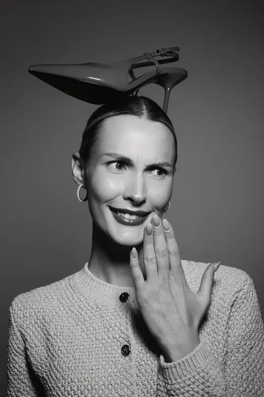 White woman with fair skin smiling, holding hand near face, balancing high heel shoe on head in studio. Concept of fashion humor and beauty.