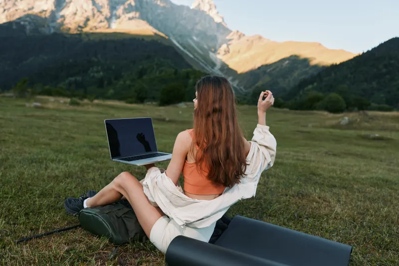 A woman sits on a grassy field with a laptop, enjoying outdoor work in a mountainous landscape. She is casual, focused on the screen, creating a calm and productive remote work scene.