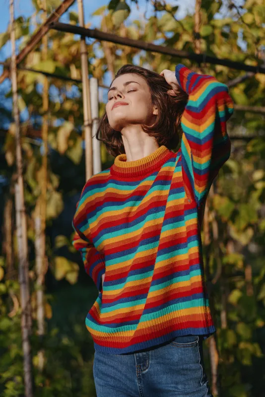 Woman in a colorful striped sweater outdoors among vines, relaxed and smiling with short hair, casual outfit, hand behind head, warm golden hour lifestyle portrait.