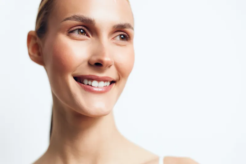 A close-up portrait of a smiling young woman with clear skin and light makeup looking away. The bright background emphasizes natural beauty and positive emotions.