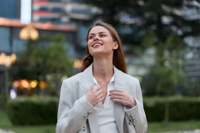 Confident business woman smiling outdoors in a stylish light blazer, showcasing modern fashion and positive emotions in an urban setting.
