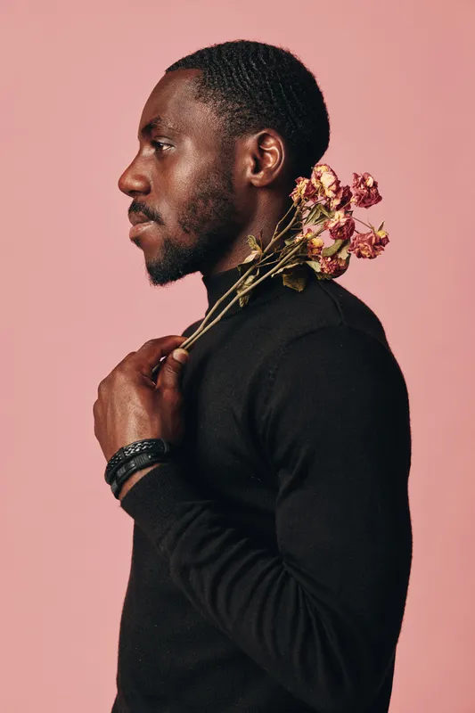 African man in black sweater holding dried flowers on shoulder in creative conceptual visual style with pink background. Portrait, profile, thoughtful expression and artistic mood.