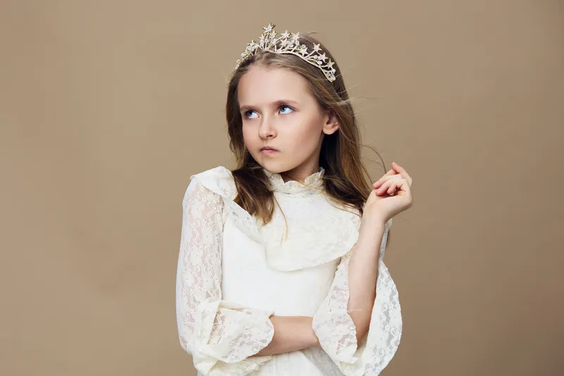 A young girl with a tiara, dressed in a white lace dress, exuding a sassy expression against a beige background, perfect for themes of childhood royalty and fashion