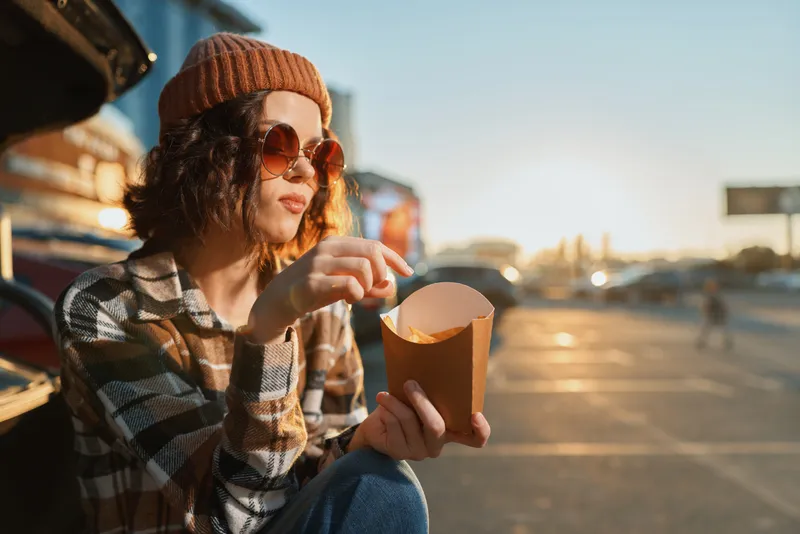 Woman with sunglasses and beanie eating fries by car in a parking lot at sunset on street, capturing authenticity, candid lifestyle, golden hour glow, mindful living and emotional storytelling.