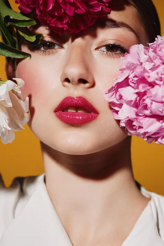 Portrait of woman with glossy red lips and flawless makeup surrounded by vibrant flowers on yellow background. Creative beauty concept focused on fashion, natural elegance, and springtime moods.