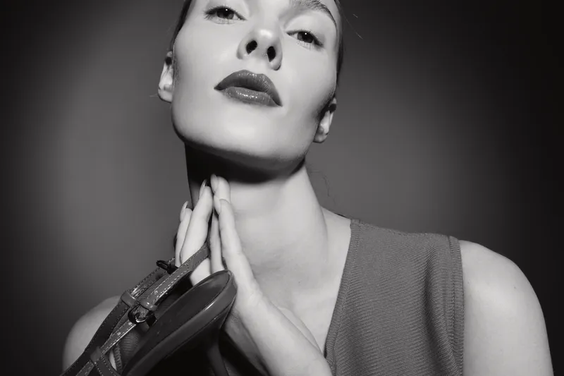 Portrait of a fair skinned Caucasian woman holding a high heel shoe near her neck in a black and white studio setting. Concept of fashion, elegance, and minimalism.