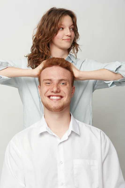 Smiling redhead man in white shirt sits with relaxed woman behind him, her hands resting on his head. Studio portrait on light background, concept of friendship and casual happiness.