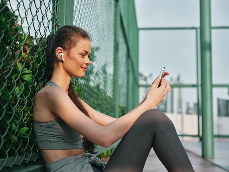 Стильная женщина в спортивной одежде наслаждается музыкой через смартфон на фоне современного города и зелёной листвы