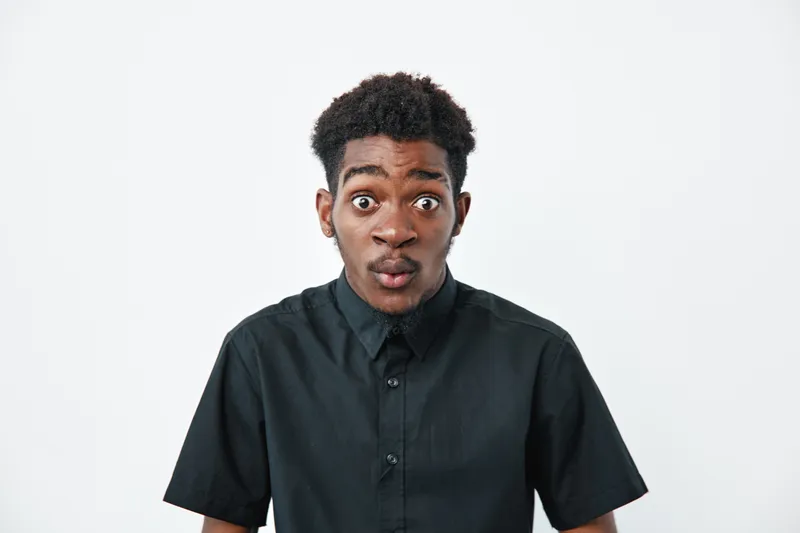 Young African American man with dark skin and curly hair wearing black shirt looking surprised with wide eyes and open mouth on white background in studio portrait.