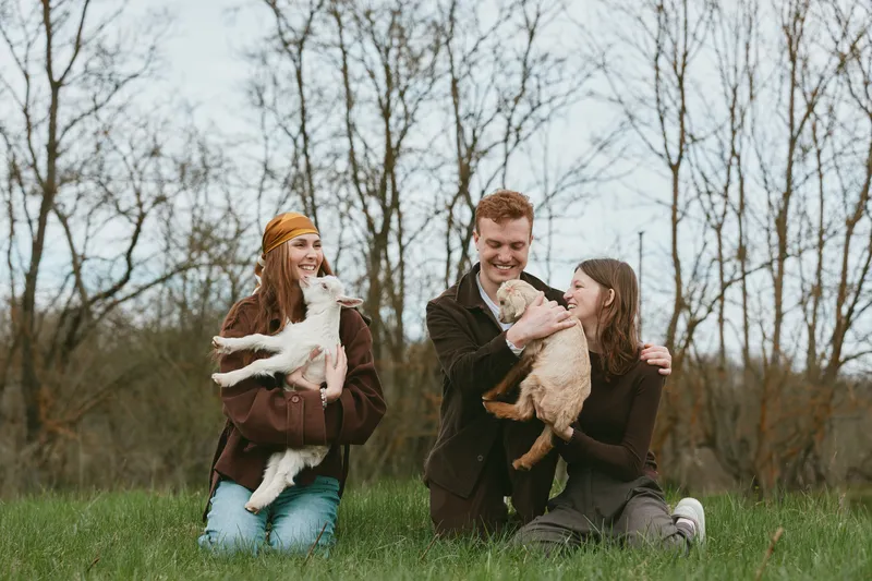 Smiling couple outdoors with two small fluffy dogs, affectionately cuddling their pets in a sunny park. Casual lifestyle moment highlighting companionship, joy, and outdoor bonding under a bright sky.