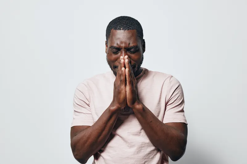 African American man with dark skin wearing light pink t shirt standing with closed eyes and hands pressed together in prayer or deep thought on white background.