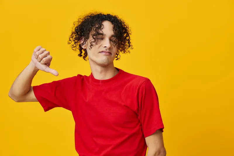 Недовольный забавный молодой человек европеоидной внешности в красной футболке показывает жест «не нравится», позируя изолированно на жёлтом студийном фоне