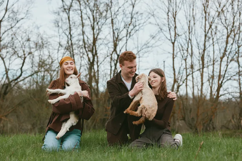 Three young adults enjoy a casual outdoor moment holding baby goats in a grassy field with bare trees in the background during a cool season.