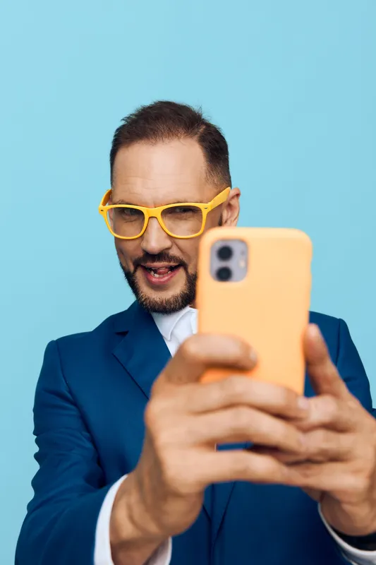 A professional man in a blue suit takes a lively selfie against a bright blue background, displaying confidence, enthusiasm and a bold, dynamic mood.