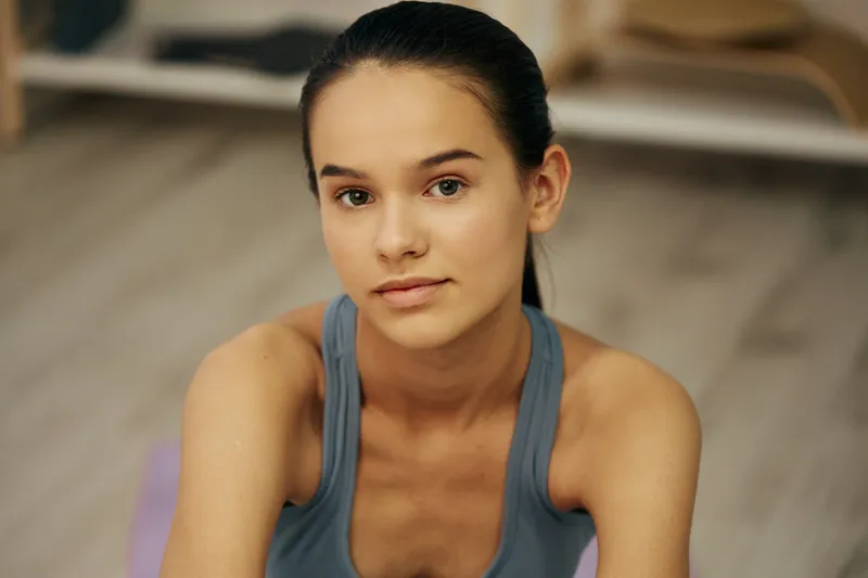 Close-up portrait of a young woman fitness model on a workout mat in a home gym, sport posture wearing a blue tank top, hair tied back, natural light, focused expression and slight sweat.