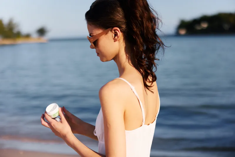 Young woman applying sunscreen by the sea in summer, wearing sunglasses and a white swimsuit, protecting skin from sun exposure during vacation.