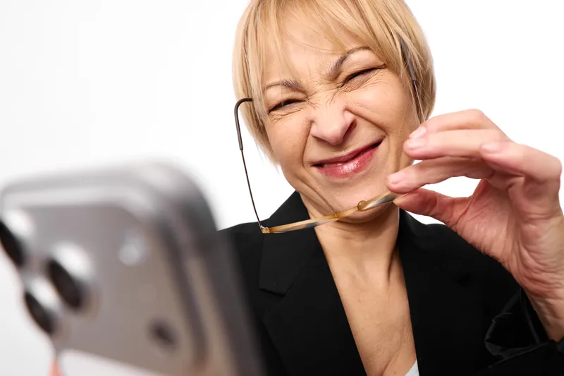Elderly woman smiles, holding eyeglasses near her face, using a smartphone indoors in a home office, relaxed posture, blurred device background, modern technology, video