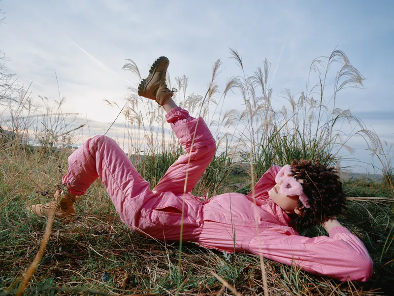 Pink outfit lying on a grassy field under a sunny sky, a carefree pose in nature. A relaxed outdoor moment with wind in hair and soft light, capturing casual fashion and a serene rural mood in the