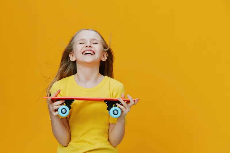 Happy girl holding skateboard, wearing yellow shirt, with joyful expression against bright yellow background, showcasing youth and energy