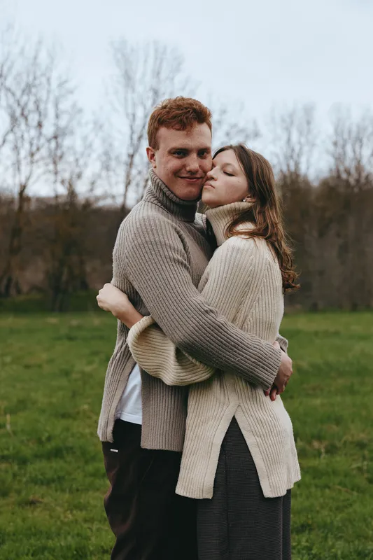 Couple embrace outdoors in cardigan and sweater, showing affection and romance in a cozy lifestyle portrait. A field backdrop, warm soft light, and natural textures convey warmth, comfort, and a