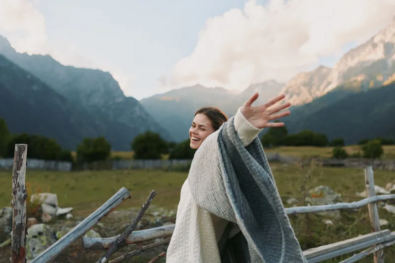 Cheerful woman outdoors, arms open, smiling amid mountains and landscape, embracing lifestyle, blanket wrapped, joyful nature, daylight and gentle breeze