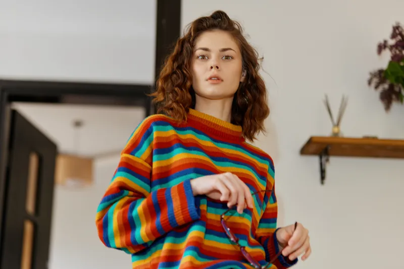 A young woman wearing a bright striped sweater stands indoors, adjusting her sleeve with a calm, focused look to convey a relaxed home lifestyle.